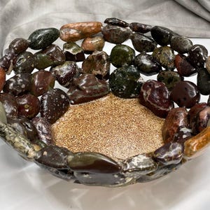 May include: A decorative bowl with a central gold-colored, textured surface, surrounded by a border of polished, multi-colored stones. The stones are various shades of brown, green, and tan, creating a natural, earthy aesthetic.