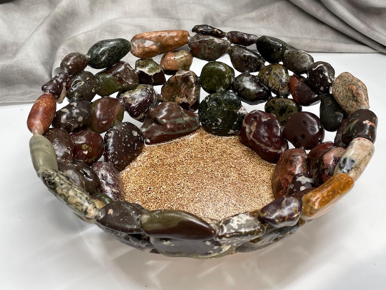 May include: A decorative bowl with a central gold-colored, textured surface, surrounded by a border of polished, multi-colored stones. The stones are various shades of brown, green, and tan, creating a natural, earthy aesthetic.