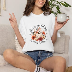 May include: A white short-sleeved t-shirt featuring a central graphic of a pumpkin surrounded by autumn-colored flowers and leaves. The text "Leaves are falling" is arched above the pumpkin, and "AUTUMN IS CALLING" is printed below in black.