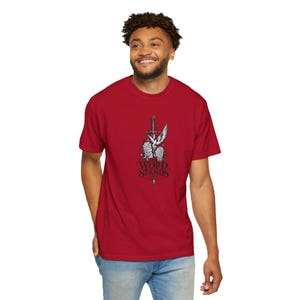 May include: A red short-sleeved crew neck t-shirt featuring a graphic print of a silver sword, a white dove, and stone tablets. Below the illustration, the text reads THE WORD STANDS in a black, stylized, serif font.