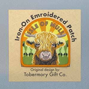 May include: An iron-on embroidered patch featuring a Highland cow with the text "Isle of Mull." The patch has an orange border and a background with a sunburst design. The text "Original design by: Tobermory Gift Co." is below the patch.