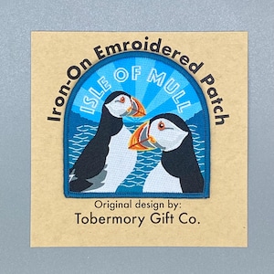 Puede incluir: Parche bordado para planchar con dos frailecillos con el texto "Isle of Mull" en un círculo azul y blanco. El parche está sobre un fondo marrón con el texto "Original design by: Tobermory Gift Co."