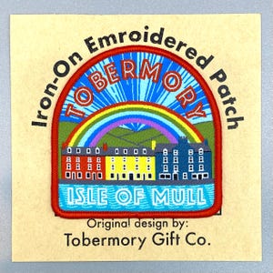 May include: An embroidered iron-on patch featuring a colorful scene of Tobermory on the Isle of Mull. The design shows a row of red, yellow, and blue buildings under a rainbow and blue sky. The text reads Iron-On Embroidered Patch, TOBERMORY, ISLE OF MULL, and Original design by: Tobermory Gift Co.