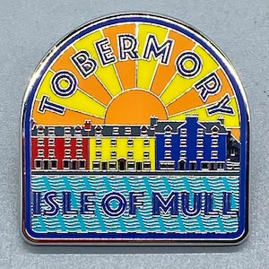 Pode incluir: Um alfinete de esmalte metálico com uma ilustração colorida de Tobermory na Ilha de Mull. O design mostra uma fila de edifícios vermelhos, amarelos e azuis contra um fundo de sol laranja e amarelo, com o texto TOBERMORY e ISLE OF MULL.