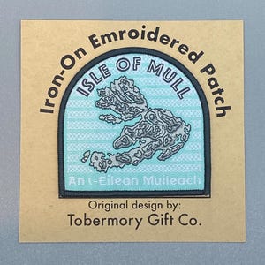Könnte beinhalten: Aufbügelbarer Stickpatch mit einer Karte der Isle of Mull, Schottland. Der Patch ist hellblau mit einem grauen Umriss der Insel. Der Text "Isle of Mull" ist in einem schwarzen Bogen über der Karte geschrieben. Der Text "An t-Eilean Muileach" ist in Schwarz unter der Karte geschrieben. Der Text "Iron-On Embroidered Patch" ist in einem schwarzen Bogen um die Oberseite des Patches geschrieben. Der Text "Original design by: Tobermory Gift Co." ist in Schwarz unter dem Patch geschrieben.
