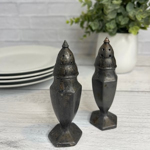 May include: A set of two vintage silver salt and pepper shakers with a hexagonal base and a domed top. The shakers have a dark, aged finish and are made of metal.