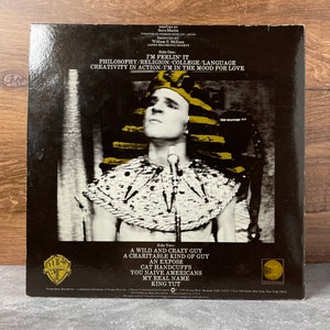 May include: A black and white photo of Steve Martin dressed as King Tut on the back of a vinyl record. The record is titled "I'm Feelin' It" and lists the songs "A Wild and Crazy Guy", "A Charitable Kind of Guy", "An Expose", "Cat Handcuffs", "You Naive Americans", "My Real Name", and "King Tut".