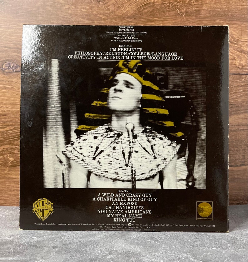 May include: A black and white photo of Steve Martin dressed as King Tut on the back of a vinyl record. The record is titled "I'm Feelin' It" and lists the songs "A Wild and Crazy Guy", "A Charitable Kind of Guy", "An Expose", "Cat Handcuffs", "You Naive Americans", "My Real Name", and "King Tut".