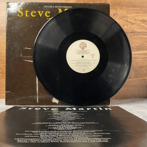 May include: A black vinyl record with a white label that reads "Steve Martin - A Wild and Crazy Guy" on the front. The back of the record jacket is black with white text that reads "Steve Martin - A Wild and Crazy Guy" and lists the musicians and recording information.