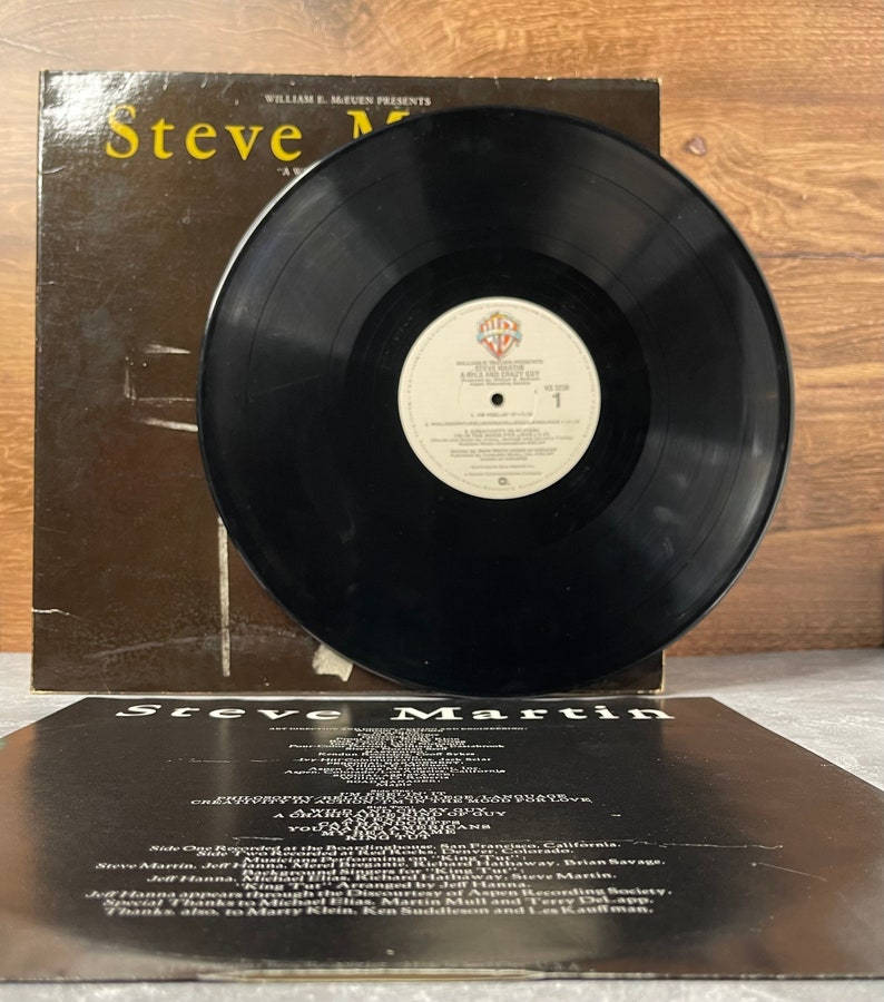 May include: A black vinyl record with a white label that reads "Steve Martin - A Wild and Crazy Guy" on the front. The back of the record jacket is black with white text that reads "Steve Martin - A Wild and Crazy Guy" and lists the musicians and recording information.