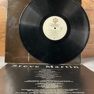 May include: A black vinyl record with a white label. The label has the text "William E. Meuen Presents Steve Martin A Wild and Crazy Guy" and the number "2". The back of the record jacket has the text "Steve Martin" and a list of credits.