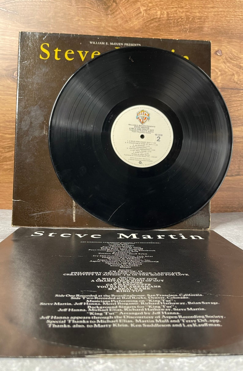 May include: A black vinyl record with a white label. The label has the text "William E. Meuen Presents Steve Martin A Wild and Crazy Guy" and the number "2". The back of the record jacket has the text "Steve Martin" and a list of credits.