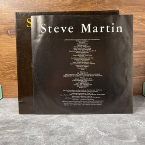 May include: A black and white record album cover with the title "Steve Martin" in white text. The album cover features a photo of Steve Martin in a black and white suit. The back of the album cover lists the album's songs, credits, and production information.