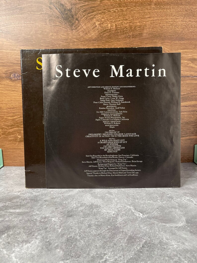 May include: A black and white record album cover with the title "Steve Martin" in white text. The album cover features a photo of Steve Martin in a black and white suit. The back of the album cover lists the album's songs, credits, and production information.