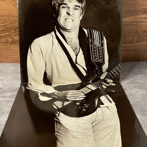 May include: Black and white photo of a man playing an electric guitar. He is wearing a white shirt with a black and white patterned design and a studded guitar strap. The photo is from a record album.
