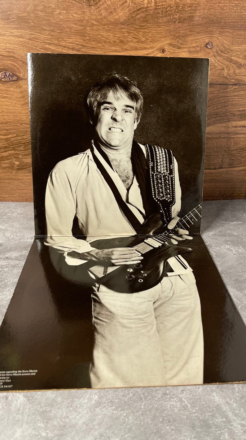 May include: Black and white photo of a man playing an electric guitar. He is wearing a white shirt with a black and white patterned design and a studded guitar strap. The photo is from a record album.