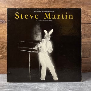 May include: A black and white photo of comedian Steve Martin wearing a white suit and bunny ears. The text "William E. McEuen Presents Steve Martin "A Wild and Crazy Guy"" is printed on the cover of the record album.