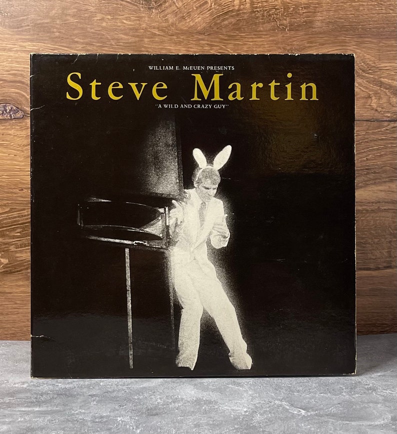 May include: A black and white photo of comedian Steve Martin wearing a white suit and bunny ears. The text "William E. McEuen Presents Steve Martin "A Wild and Crazy Guy"" is printed on the cover of the record album.
