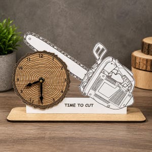 Könnte beinhalten: Eine Holzuhr mit einem Zifferblatt in Form eines Baumstamms und schwarzen Zeigern, kombiniert mit einem Kettensägen-Design. Die Uhr steht auf einem Holzsockel mit dem Text "TIME TO CUT".