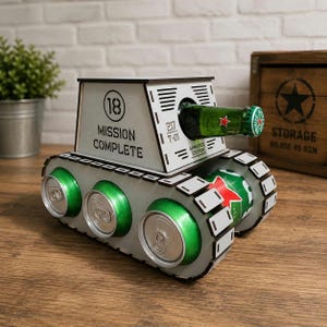 Puede incluir: Un soporte para bebidas de madera blanca con forma de tanque que presenta el texto 18 MISSION COMPLETE y 217 T-01 ARMORED EDITION. La estructura incorpora latas de bebida verdes como ruedas y una botella de vidrio verde como cañón.