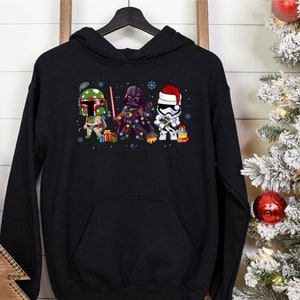 May include: Black hooded sweatshirt with a Christmas-themed graphic of Star Wars characters: Boba Fett, Darth Vader, and a Stormtrooper. The design incorporates festive elements like fairy lights, presents, and snowflakes, ideal for the holiday period.