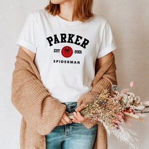 May include: White t-shirt with black text that reads "Parker Est. 2001 Spiderman". The text is in a collegiate font style. A red circle with a black spider logo is above the word "Spiderman".