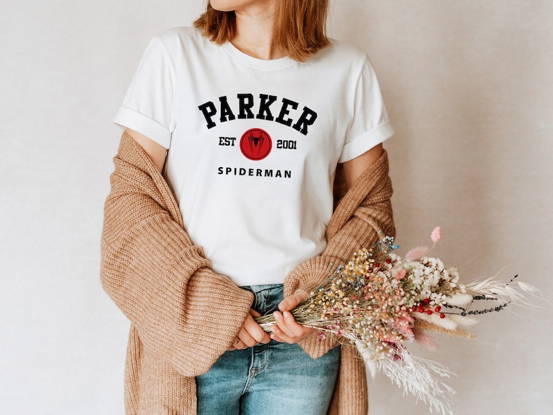 May include: White t-shirt with black text that reads "Parker Est. 2001 Spiderman". The text is in a collegiate font style. A red circle with a black spider logo is above the word "Spiderman".