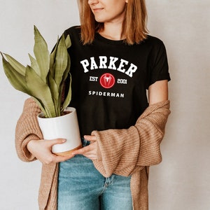 May include: Black t-shirt with white text that reads "Parker Est. 2001 Spiderman". The text is in a collegiate font. A red spider logo is above the word "Spiderman".