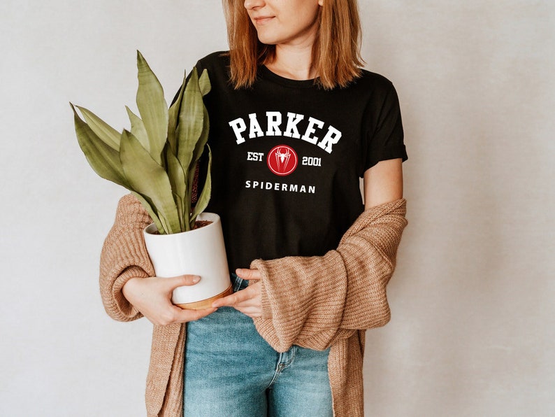 May include: Black t-shirt with white text that reads "Parker Est. 2001 Spiderman". The text is in a collegiate font. A red spider logo is above the word "Spiderman".