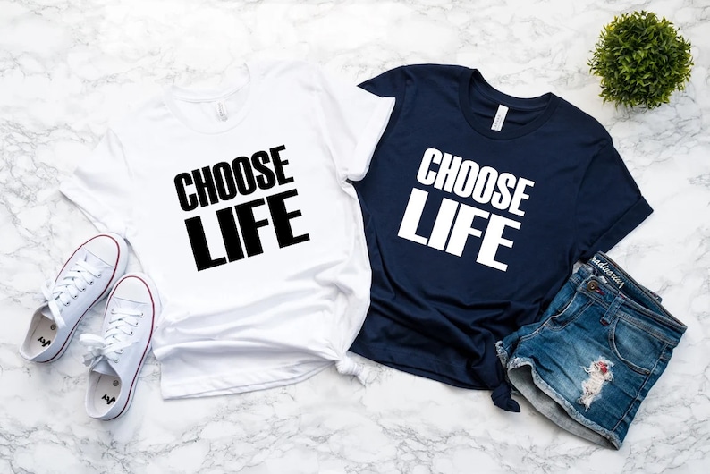Puede incluir: Dos camisetas, una blanca y otra azul marino, cada una con las palabras "CHOOSE LIFE" en negrita, en letras blancas y negras. Tambi&eacute;n se ven zapatillas blancas y pantalones cortos vaqueros. Las camisetas probablemente forman parte de un conjunto a juego.