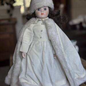 May include: A porcelain doll in a white dress, fur-trimmed cape, and a fur hat. The doll has brown hair, blue eyes, and a painted face. The dress is buttoned with pearl beads. The doll is on a wooden surface. The cape and hat have a soft, fluffy texture.