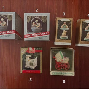 May include: A collection of Christmas ornaments in their original boxes. The ornaments include a snow globe, a baby crib, and a baby carriage. The boxes have text that reads "Light! Motion! Music!" and "Baby's First Christmas."