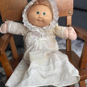 Op de afbeelding: Een vintage Cabbage Patch Kid-pop met een rond gezicht, groene ogen en lichtbruin haar. De pop draagt een witte jurk met roze bloemenaccenten en een met kant afgezette muts. Ze zit in een houten schommelstoel.