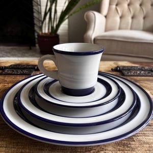 Puede incluir: Un juego de taza y platillo de cerámica blanca apilados sobre dos platos de cerámica blanca más grandes. Cada pieza presenta un borde azul oscuro liso y una base a juego, creando una apariencia uniforme y superpuesta para este conjunto.
