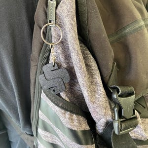 May include: A black zipper pull with the word "adidas" printed on it is attached to a silver keyring with a black leather keychain shaped like the letter "S". The keychain is attached to a grey and black backpack.