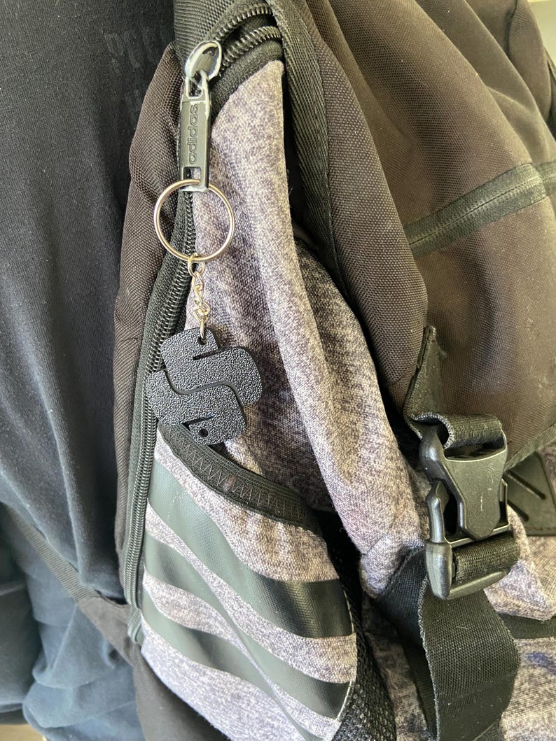 May include: A black zipper pull with the word "adidas" printed on it is attached to a silver keyring with a black leather keychain shaped like the letter "S". The keychain is attached to a grey and black backpack.