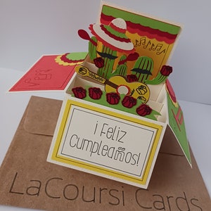 May include: A pop-up card with a 3D design of two cacti wearing sombreros and playing a guitar. The card is decorated with red roses and musical notes. The card reads "&iexcl;Feliz cumplea&ntilde;os!" which means "Happy Birthday!" in Spanish.