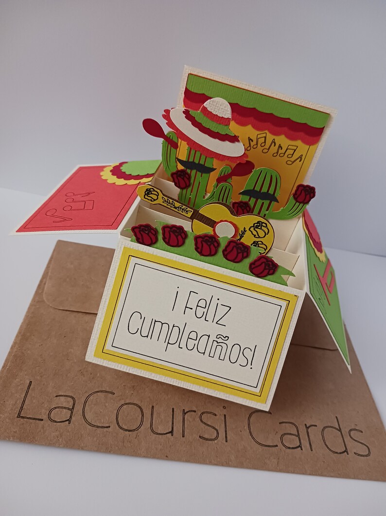 May include: A pop-up card with a 3D design of two cacti wearing sombreros and playing a guitar. The card is decorated with red roses and musical notes. The card reads "&iexcl;Feliz cumplea&ntilde;os!" which means "Happy Birthday!" in Spanish.