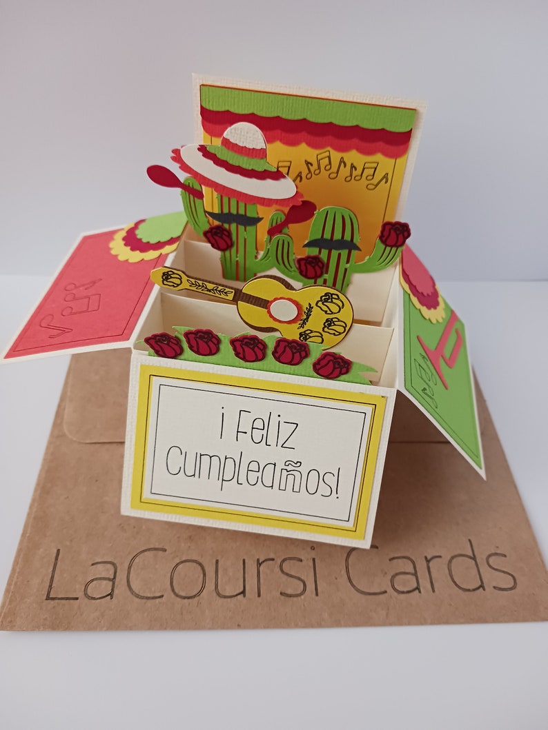May include: A pop-up card with a cactus and sombrero design. The card is open and shows a guitar and the words "&iexcl;Feliz Cumplea&ntilde;os!" in Spanish.