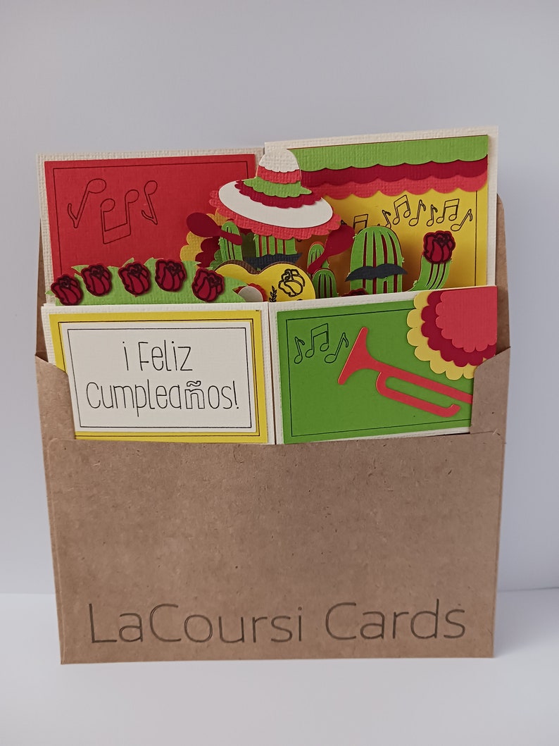 May include: A brown paper envelope with a pop-up card inside. The card features a colorful Mexican fiesta theme with a sombrero, cacti, musical notes, and the text "&iexcl;Feliz Cumplea&ntilde;os!" The envelope has the text "LaCoursi Cards" printed on the front.