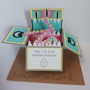 May include: A pop-up greeting card featuring a monster design with pink tentacles and three eyes. The card is open, revealing a twenty-sided die inside. The front panel displays the text "Add +1 to your wisdom modifier" above a lock icon.