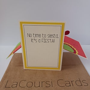 May include: A yellow and white card with the text "No time to siesta, it's a FIESTA!" on the front. The card is folded in half and has a brown envelope with the text "LaCoursi Cards" on the front.