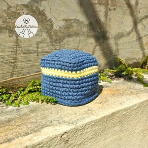 May include: A crocheted blue hat with a yellow stripe. The hat is handmade and has a rounded top and a flat brim. The hat is sitting on a concrete surface with green plants in the background. The hat is a small size.