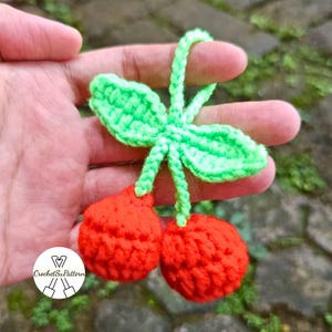 Peut inclure: Une cerise au crochet faite à la main, avec deux sphères rouges et des feuilles et une tige vertes. Le travail au crochet est détaillé, avec des points visibles. La cerise est tenue dans une main, avec un arrière-plan flou.