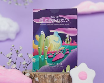 May include: A purple box with the title "Oráculo El Despertar" and the text "Sanación y afirmaciones de amor propio". The box features a colorful illustration of a person sitting in a landscape with cacti and flowers. The text "Paulie Dahl" and "Ilustrado por Aleja Jones" is also on the box.