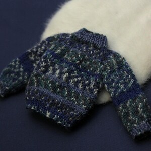 May include: A small, long-sleeved knitted jumper featuring a variegated pattern of dark blue, teal, and grey yarn. The garment has a ribbed crew neckline, ribbed cuffs, and a ribbed hem, constructed with a thick, textured knit stitch throughout.