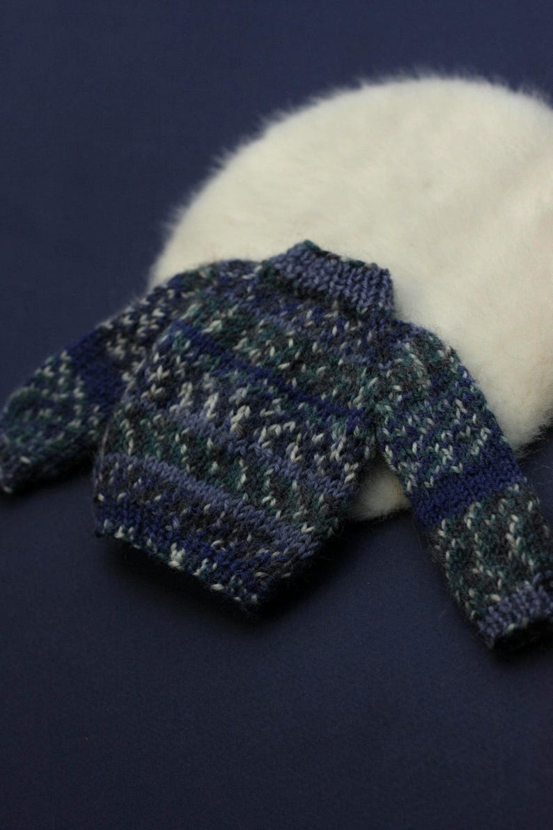 May include: A small, long-sleeved knitted jumper featuring a variegated pattern of dark blue, teal, and grey yarn. The garment has a ribbed crew neckline, ribbed cuffs, and a ribbed hem, constructed with a thick, textured knit stitch throughout.