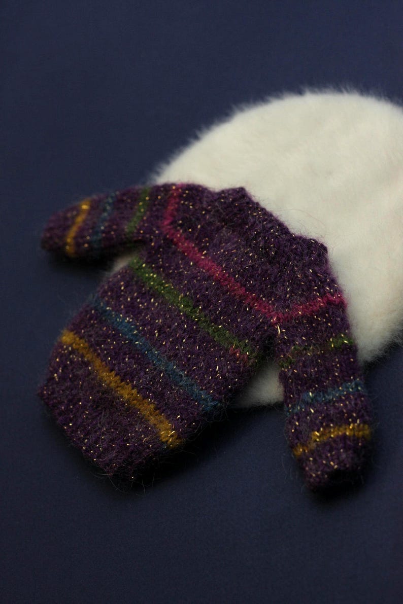 May include: A miniature long-sleeved jumper in a deep purple fuzzy knit texture with horizontal stripes in gold, green, and blue. The jumper features a crew neckline and a metallic shimmer throughout the yarn, resting against a white fuzzy circular base.