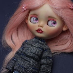 May include: A doll with long, straight pastel pink hair adorned with small pink flower clips. The doll has large, light purple eyes, pink eyelashes, and red lips. It wears a grey knitted jumper with thin blue and black stripes and blue trousers with white polka dots.