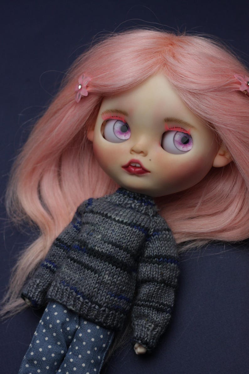 May include: A doll with long, straight pastel pink hair adorned with small pink flower clips. The doll has large, light purple eyes, pink eyelashes, and red lips. It wears a grey knitted jumper with thin blue and black stripes and blue trousers with white polka dots.